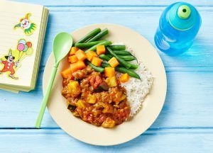 A plate of curry with rice, green beans, and cubed squash, served with a green spoon, next to a notebook and water bottle.