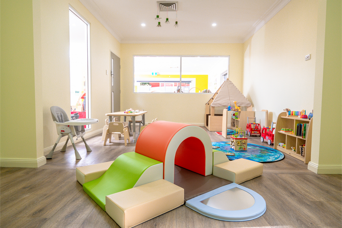 A brightly lit preschool classroom with tables, chairs, toy shelves, and a small play tunnel on a wooden floor.