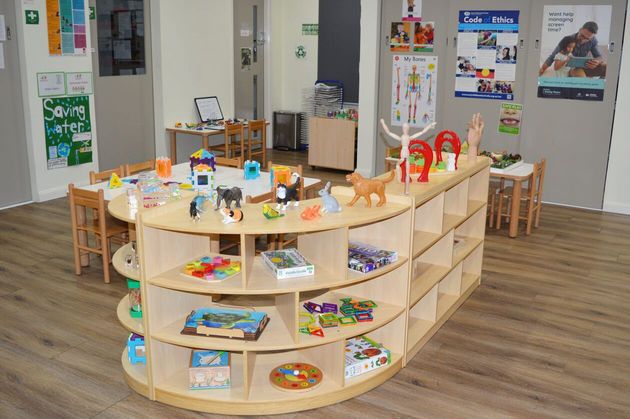 A brightly lit preschool classroom with wooden tables, chairs, and open shelving units filled with various toys.
