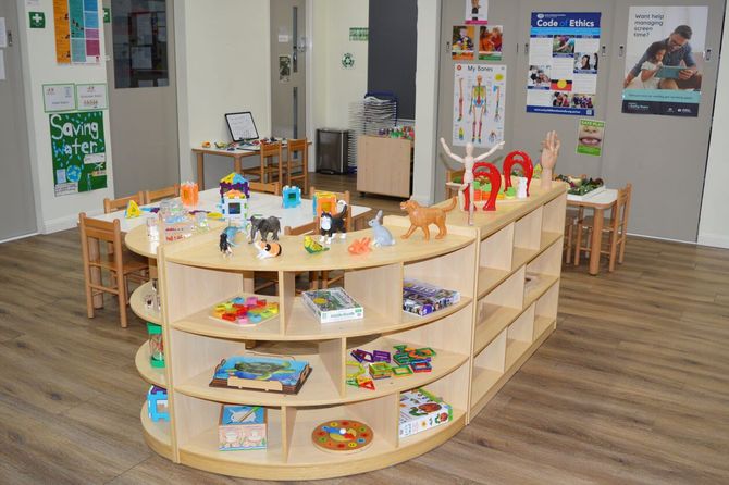 A brightly lit preschool classroom with wooden tables, chairs, and open shelving units filled with various toys.