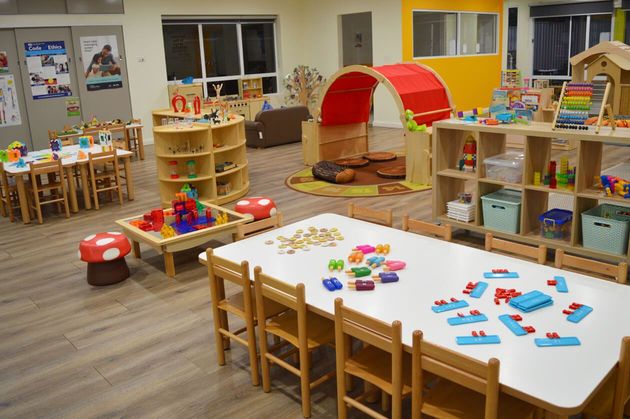 A brightly lit preschool classroom with tables, chairs, toy shelves, and a small play tunnel on a wooden floor.