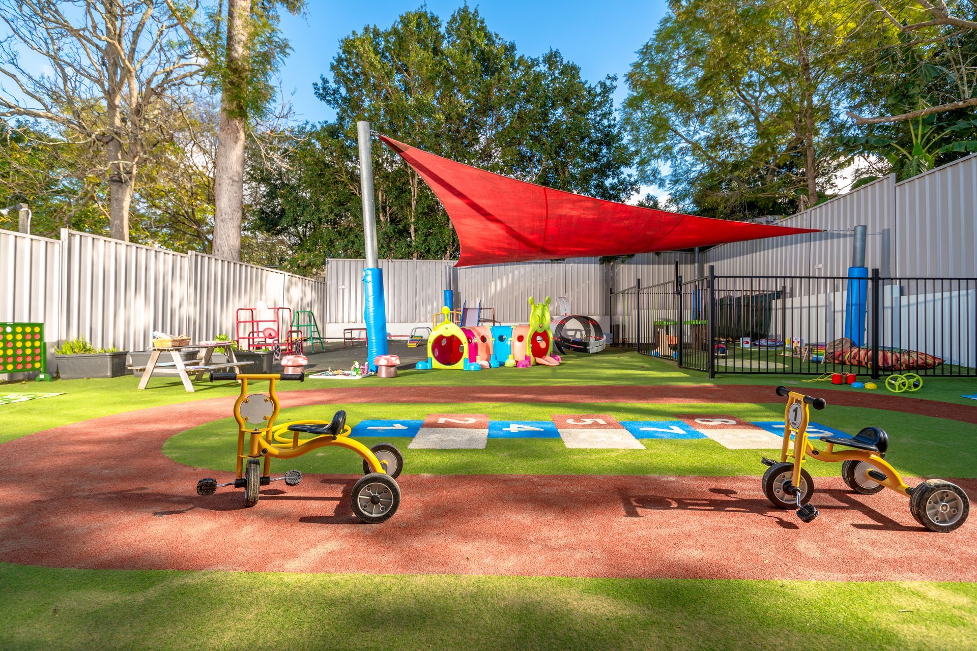 A bright, colorful preschool classroom filled with wooden tables, chairs, toys, and a red arched play structure.