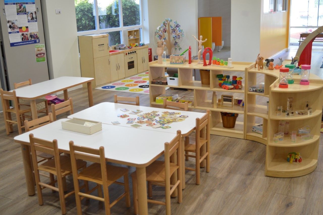 A brightly lit preschool classroom with wooden tables, chairs, and open shelving units filled with various toys.