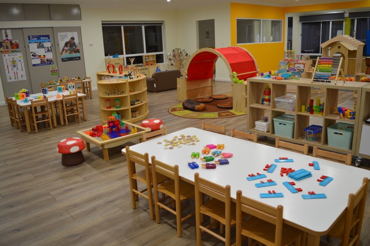 A brightly lit preschool classroom with tables, chairs, toy shelves, and a small play tunnel on a wooden floor.