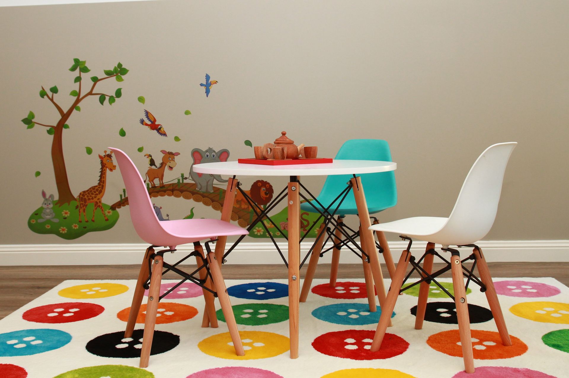 A small, white round table with three colorful chairs on a button-patterned rug in front of a jungle-themed wall mural.