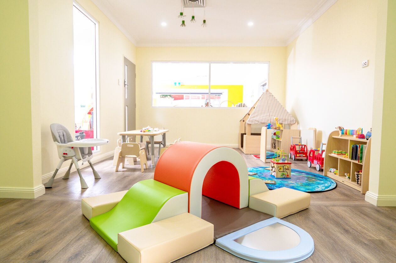 A brightly lit preschool classroom with wooden tables, chairs, and open shelving units filled with various toys.