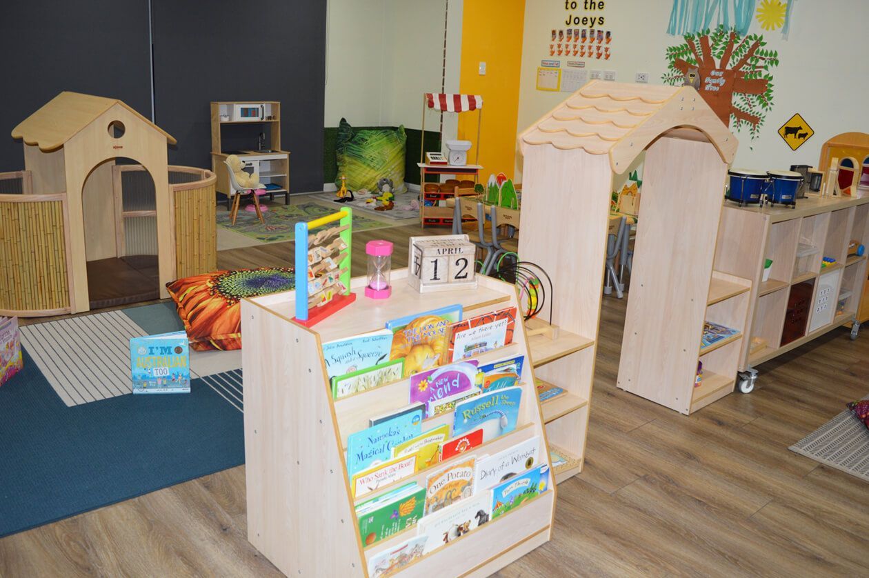 A brightly lit preschool classroom with tables, chairs, toy shelves, and a small play tunnel on a wooden floor.