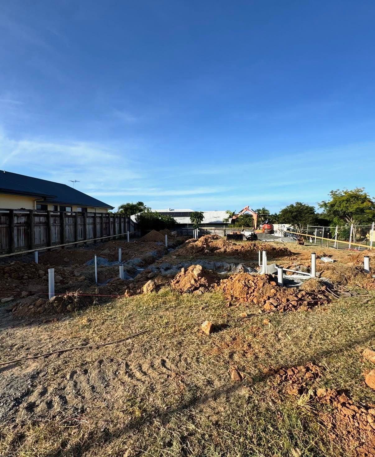 Cleared construction site with dirt ground, white posts, and a house in the distance under a blue sky