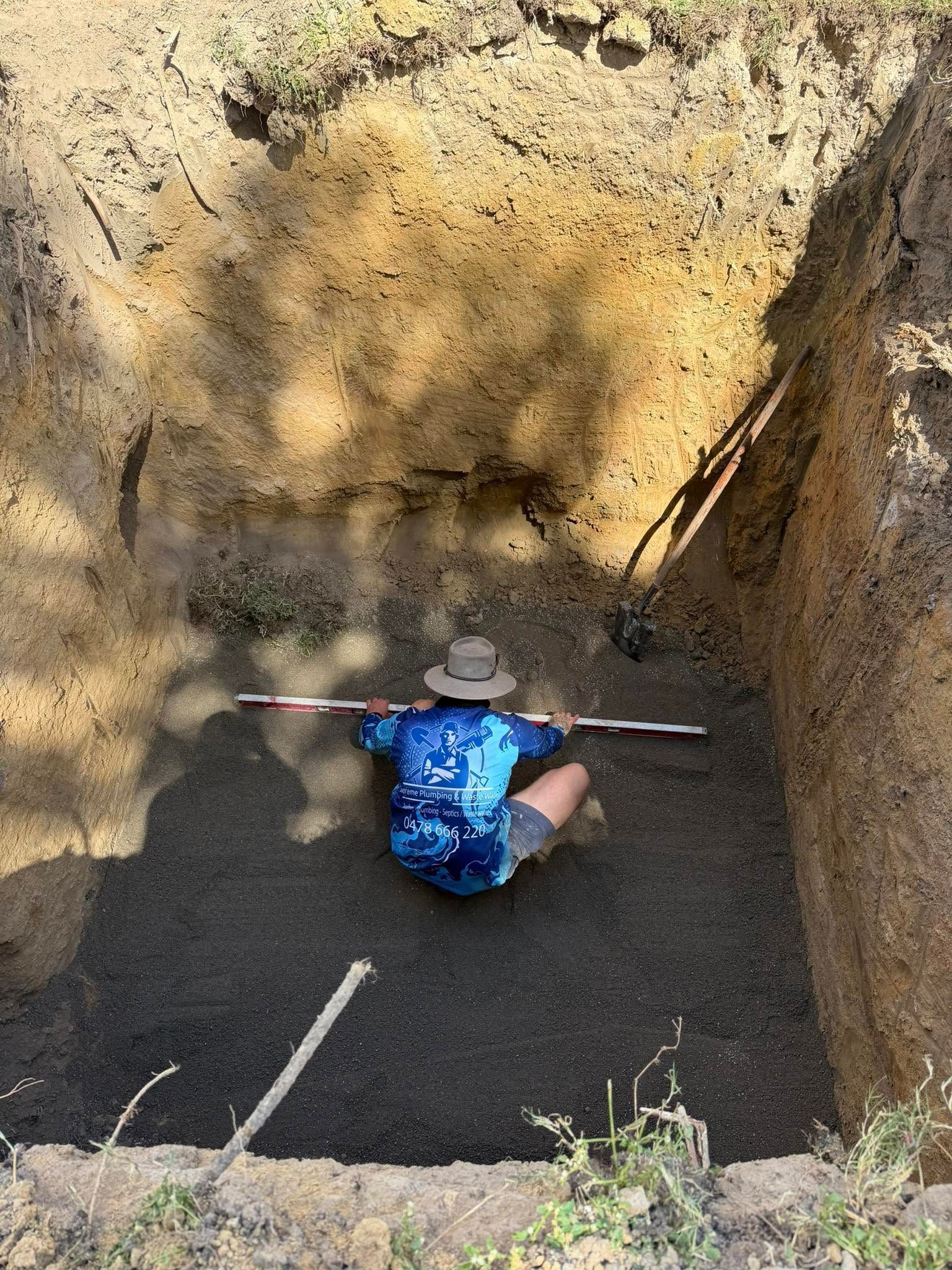Person in blue shirt sitting in a dirt excavation pit with a shovel across their shoulders