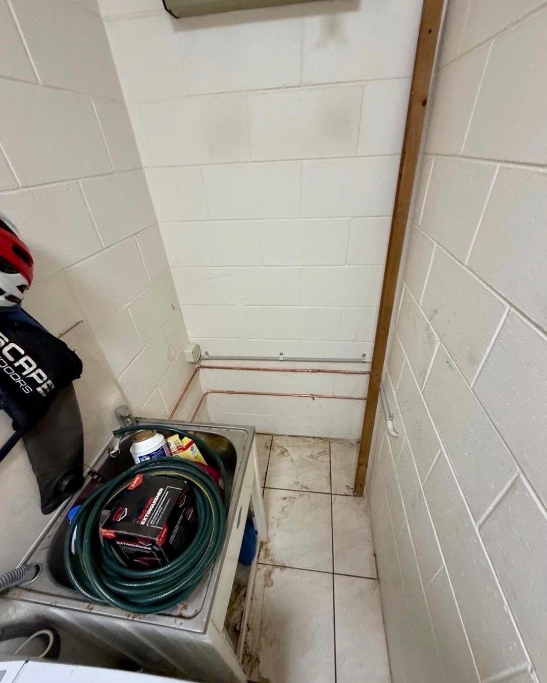 Narrow utility closet with white tile walls, pipes, and a bucket holding coiled hoses and tools.