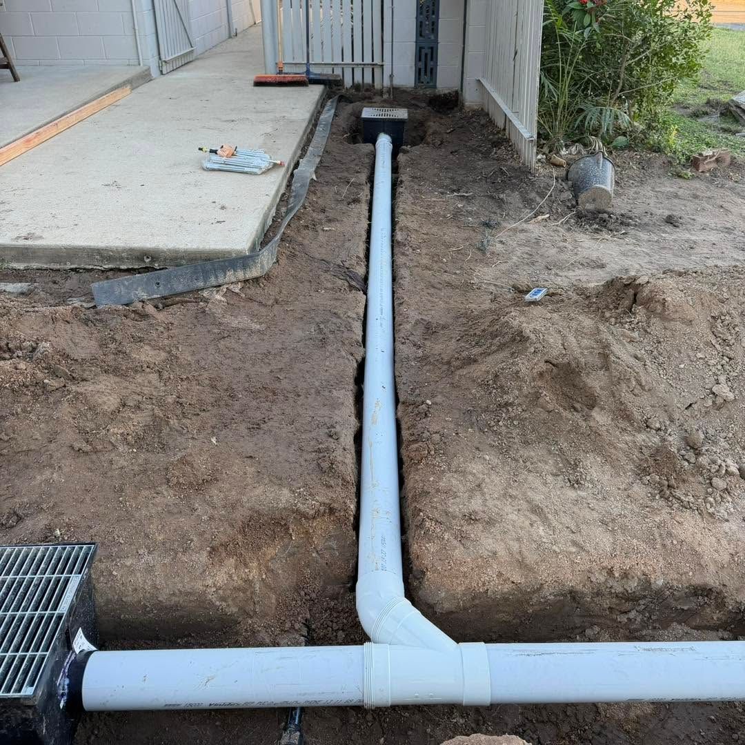Trenched white PVC pipe runs through a dirt yard beside a building and drain grate.