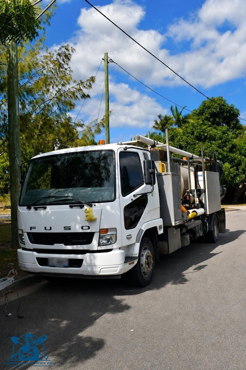 White utility truck parked on a sunny street beside a utility pole and overhead power lines.