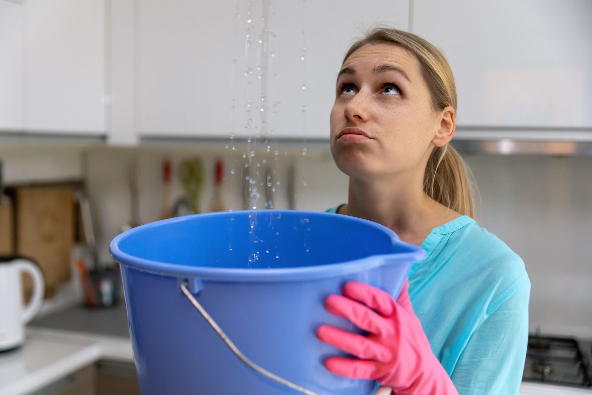 Woman in pink gloves holding a blue bucket under a dripping faucet in a kitchen