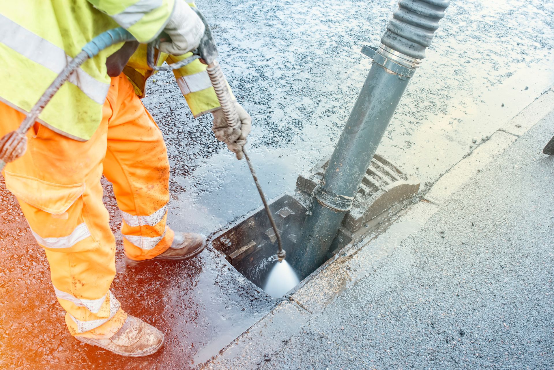 Worker in orange safety gear using a tool to inspect a street opening with a pipe and sprayed water