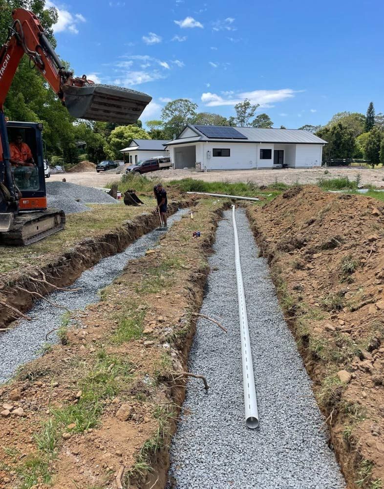 A Man is Digging a Trench in the Dirt in Front of a House — Hoges Plumbing In Kiama, NSW
