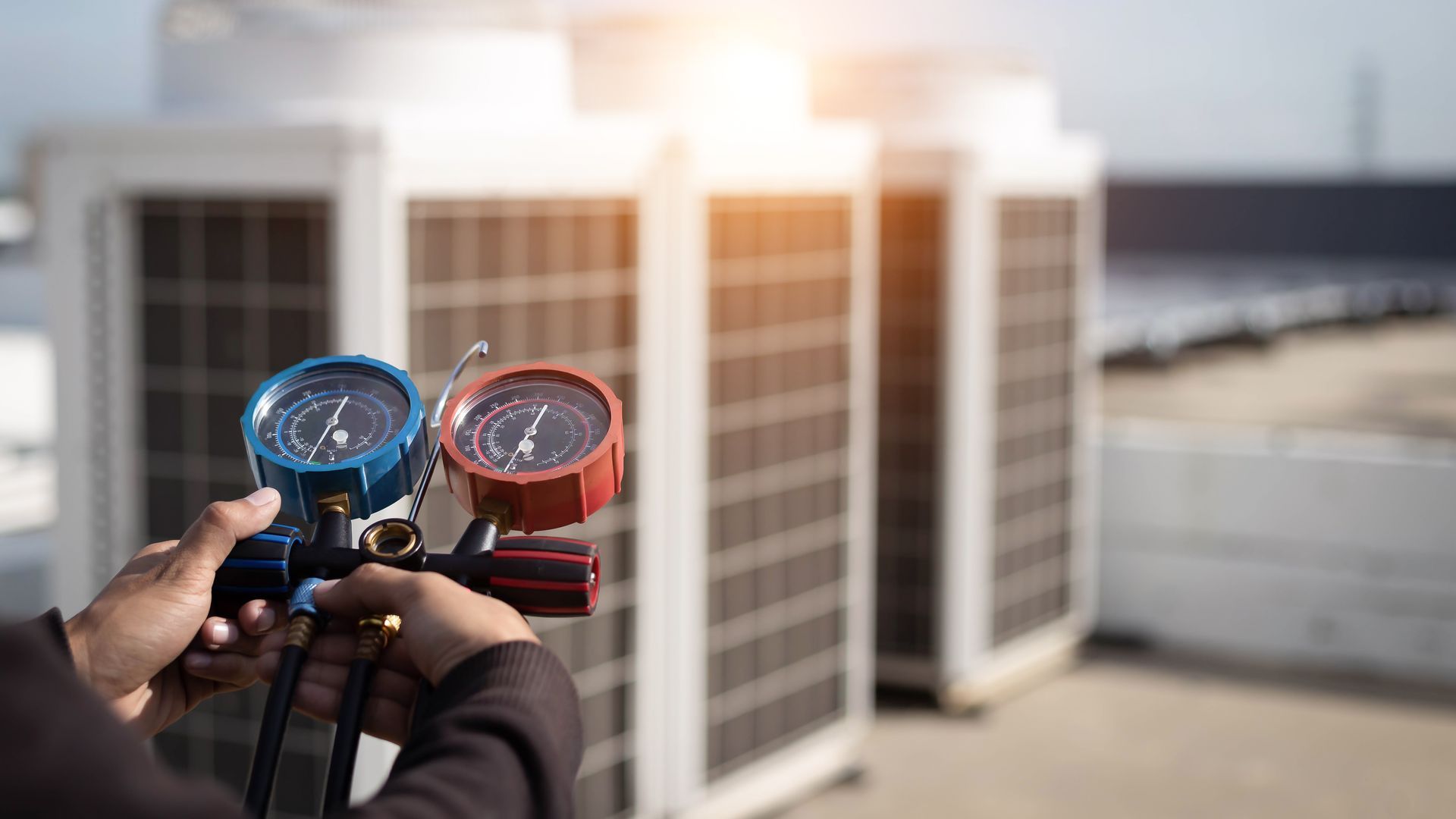 Hands holding red and blue pressure gauges in front of rooftop HVAC units at sunset