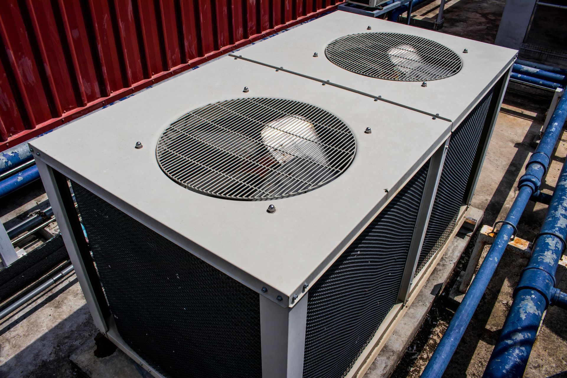 Rooftop air conditioning unit with two fans beside blue pipes and a red wall