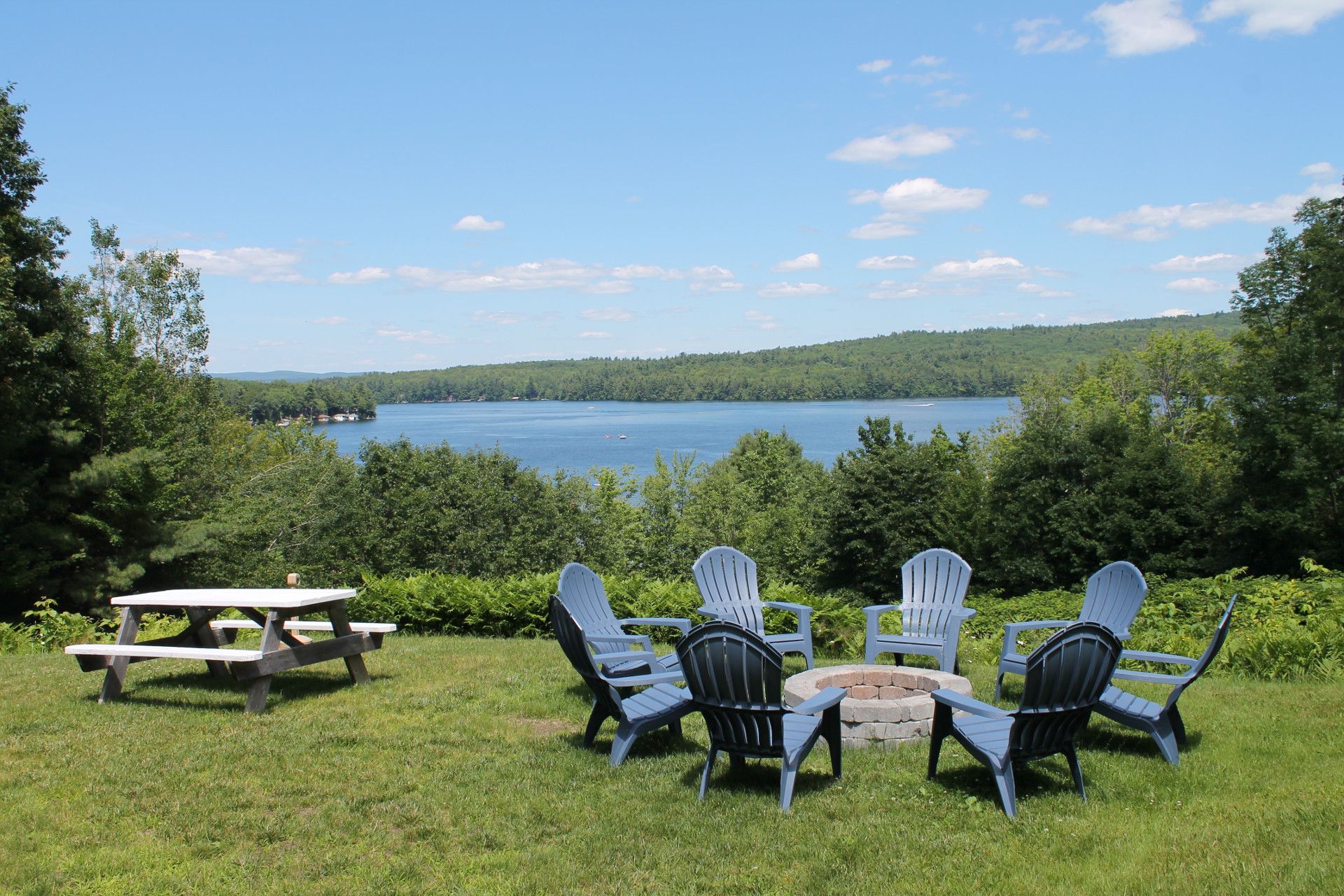 A lakeside fire pit surrounded by blue chairs, picnic table, and forest under a blue sky.