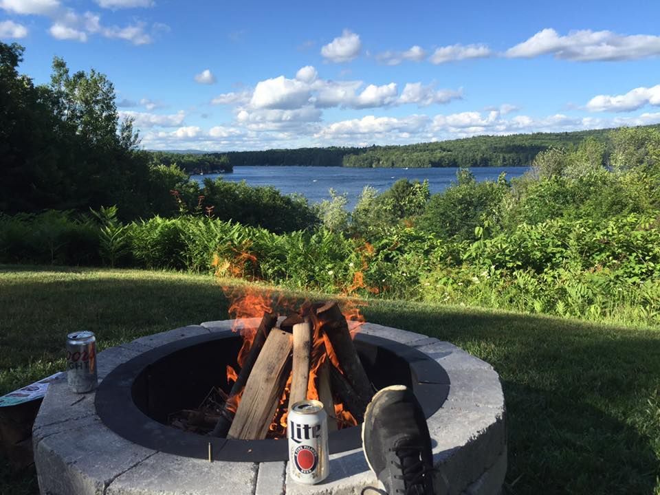 Fire pit with burning logs, overlooking a lake surrounded by green trees under a blue sky with clouds.