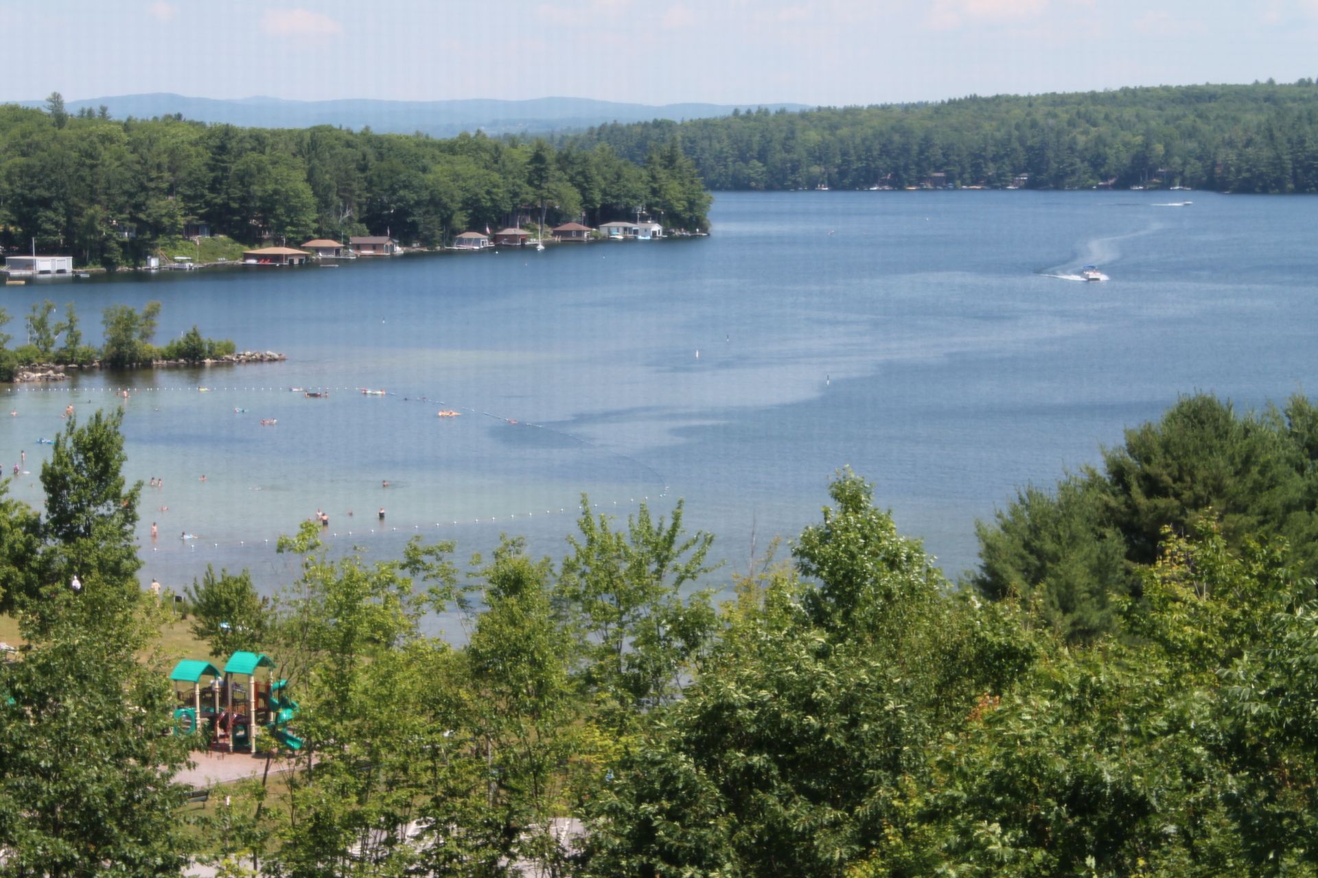 Lakeside view with green trees, calm water, and distant cabins under a blue sky.