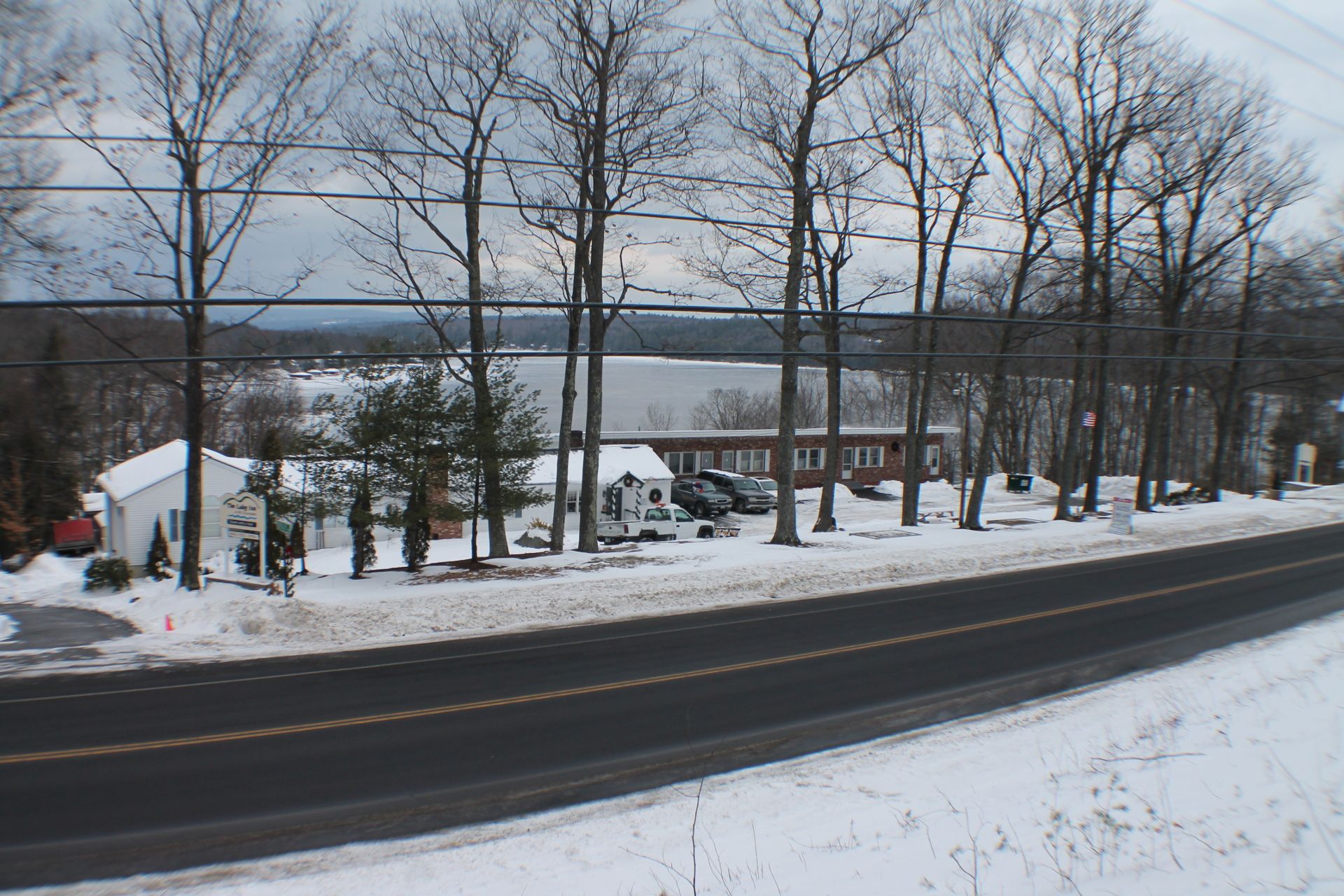 Roadside view with buildings, trees, and lake, covered in snow; overcast day.
