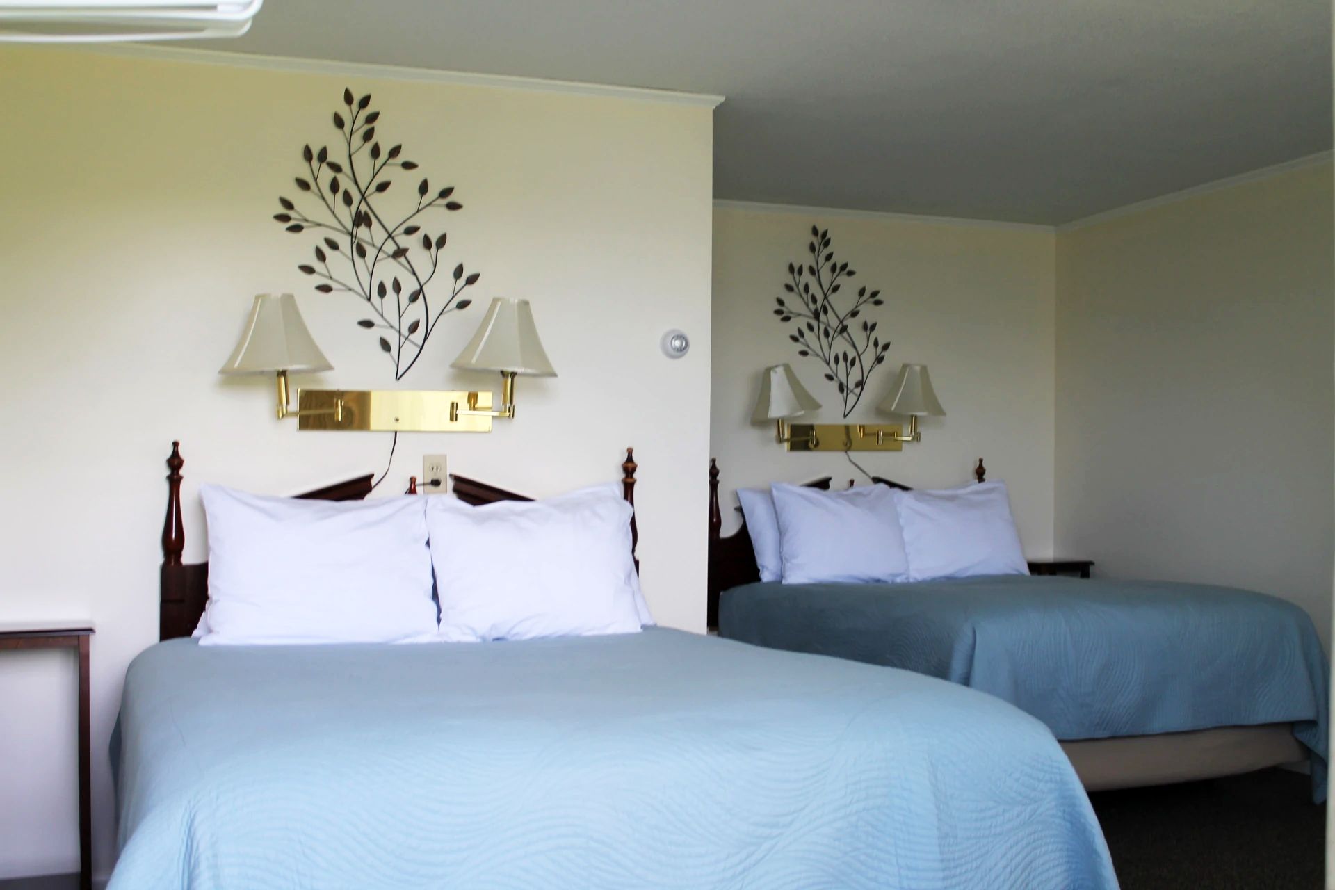 Two beds in a motel room with light blue bedspreads, floral wall art, and brass lamps.