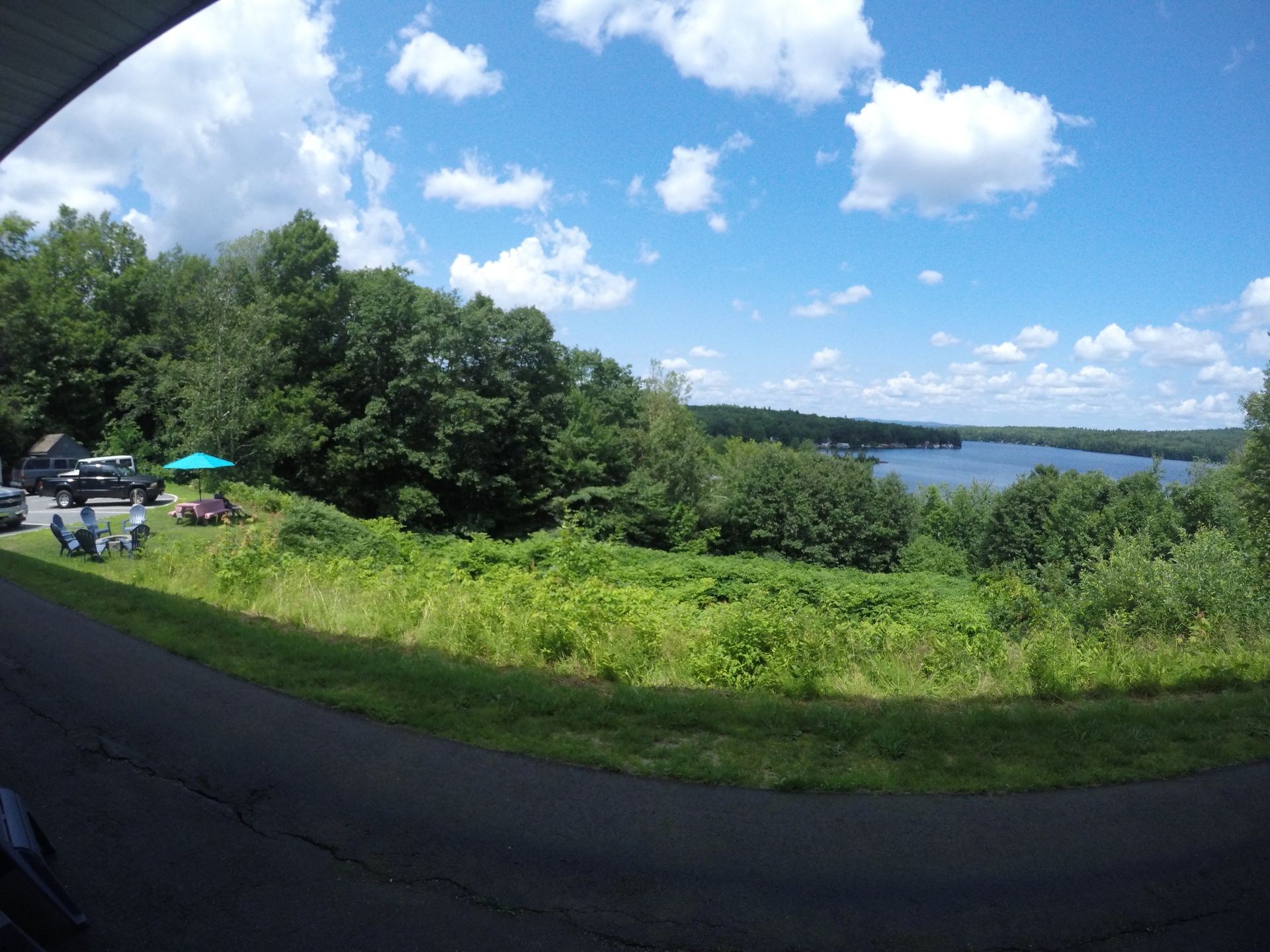 Overlook of a lake and trees under a partly cloudy sky; a few people and cars are near the trees.