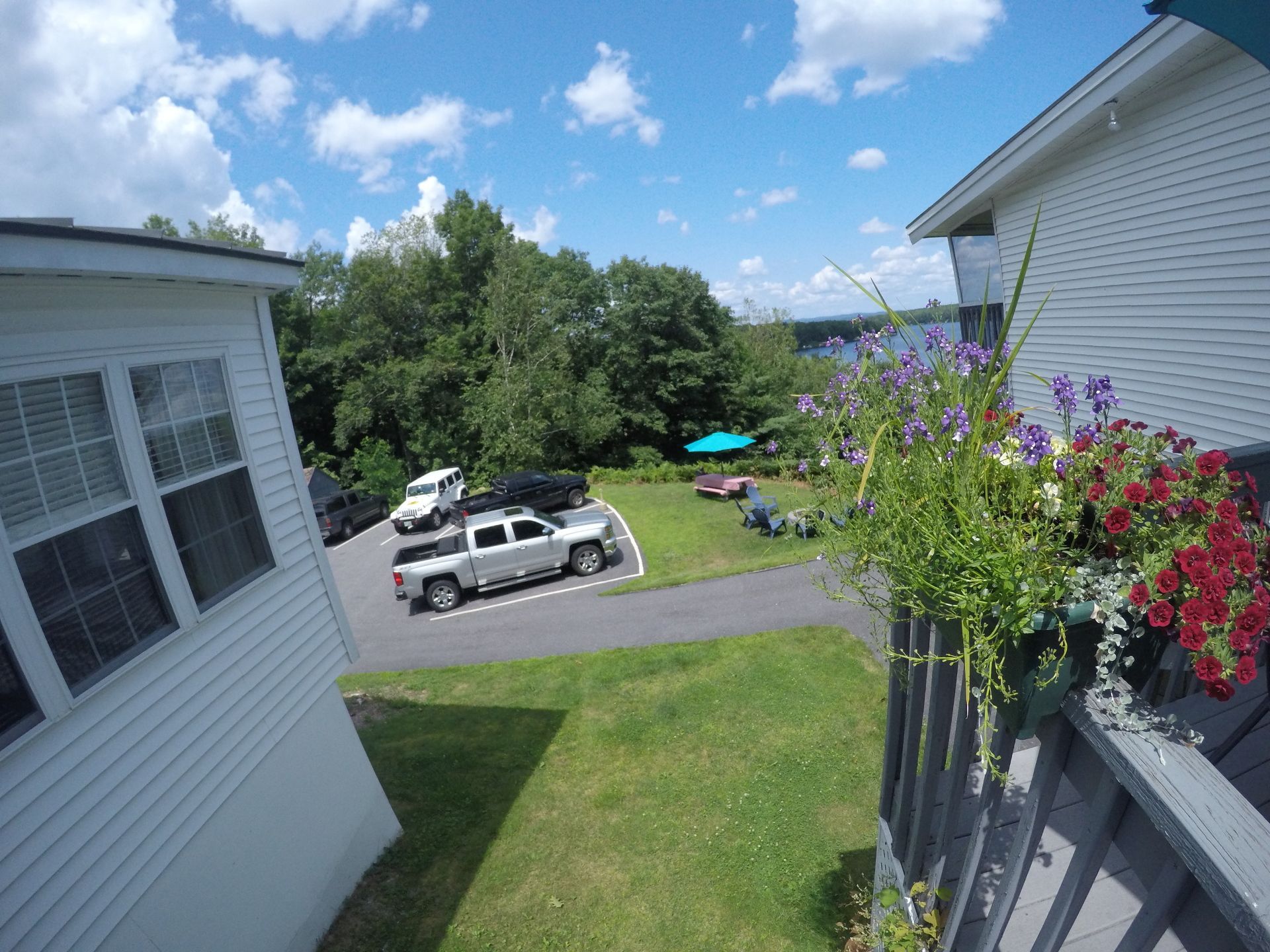 View from a porch with flowers overlooking a grassy area, a parking lot with a truck, and trees under a blue sky.