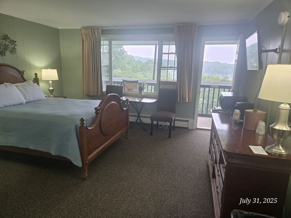 Hotel room with a bed, dresser, table, chairs, and balcony overlooking a scenic view.
