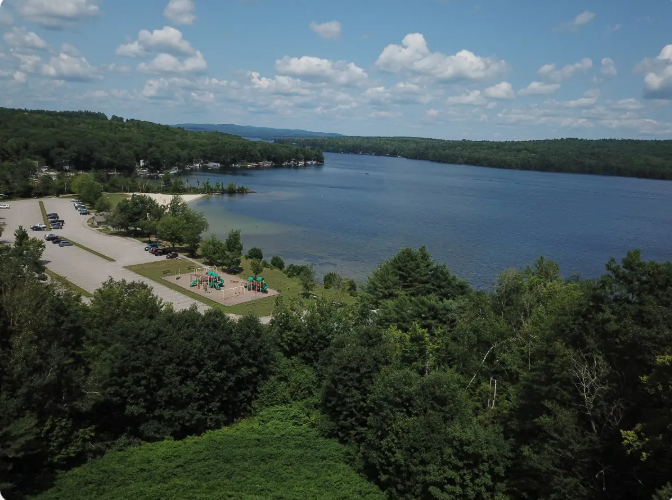 Overlooking a lake with a beach, parking lot, and playground; trees surround the water under a blue sky.