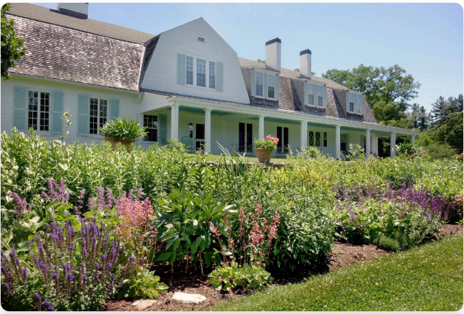 White house with a long porch, overlooking a colorful flower garden in the sun.