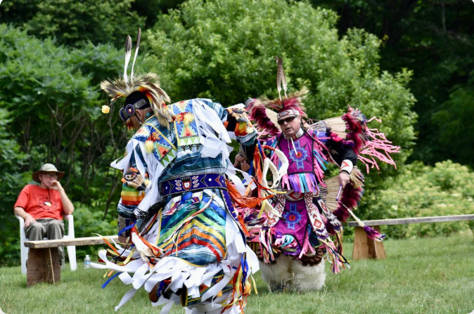 Two dancers in colorful regalia perform outdoors; a spectator watches nearby.