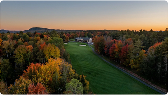 Golf course fairway lined with fall foliage, clubhouse in the distance at sunset.