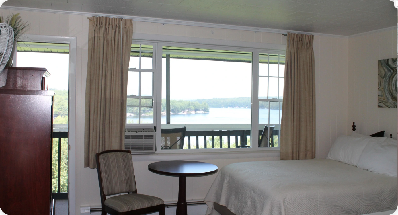Bedroom with lake view: bed, small table, chair, windows, beige curtains, and dark wood wardrobe.