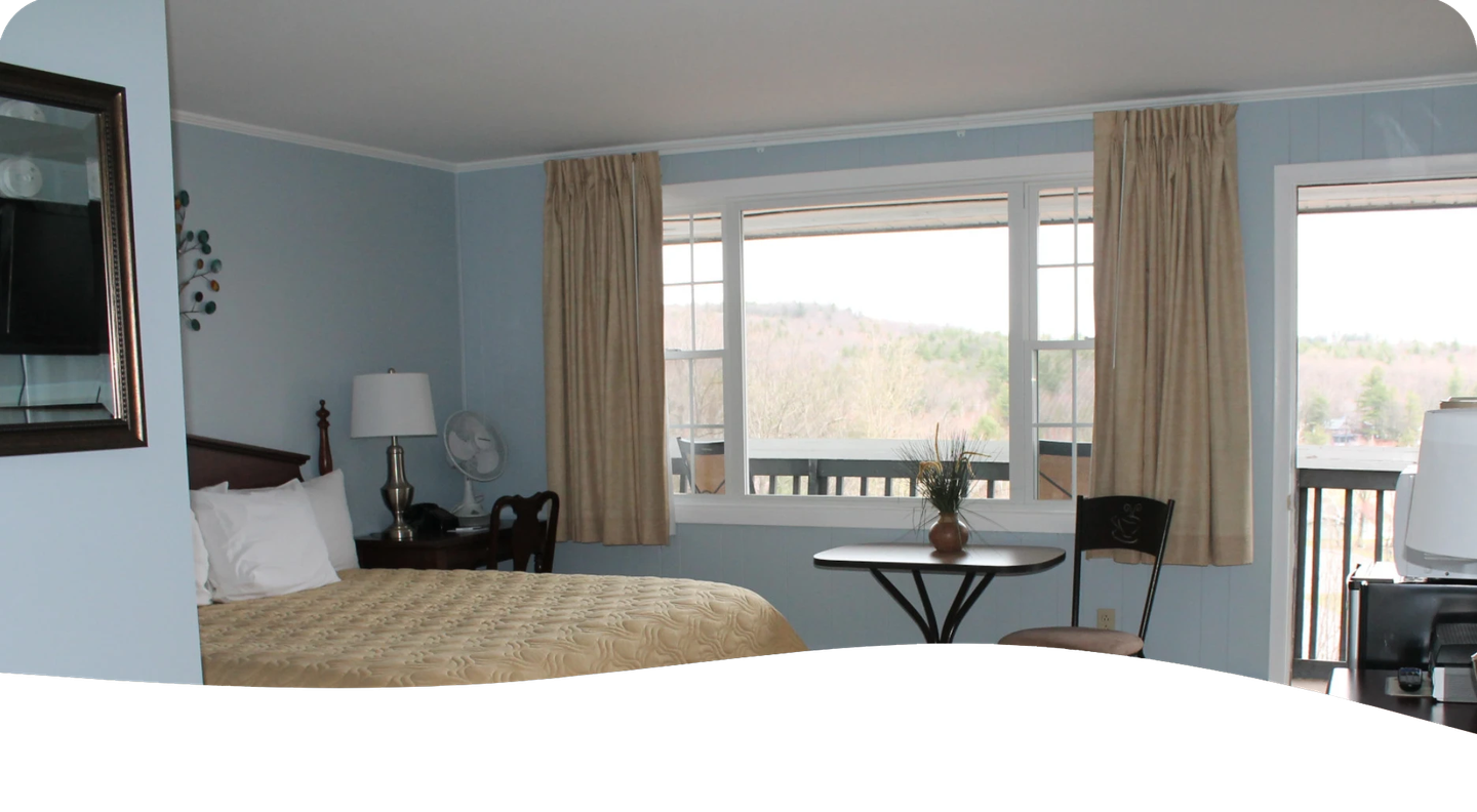 Bedroom with bed, desk, table, window overlooking trees, beige curtains.