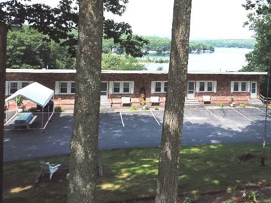 Brick motel building near a lake, seen through two trees. Black asphalt parking lot, blue sky.
