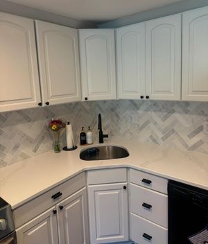 White corner kitchen with cabinets, chevron backsplash, black sink, and white countertop.