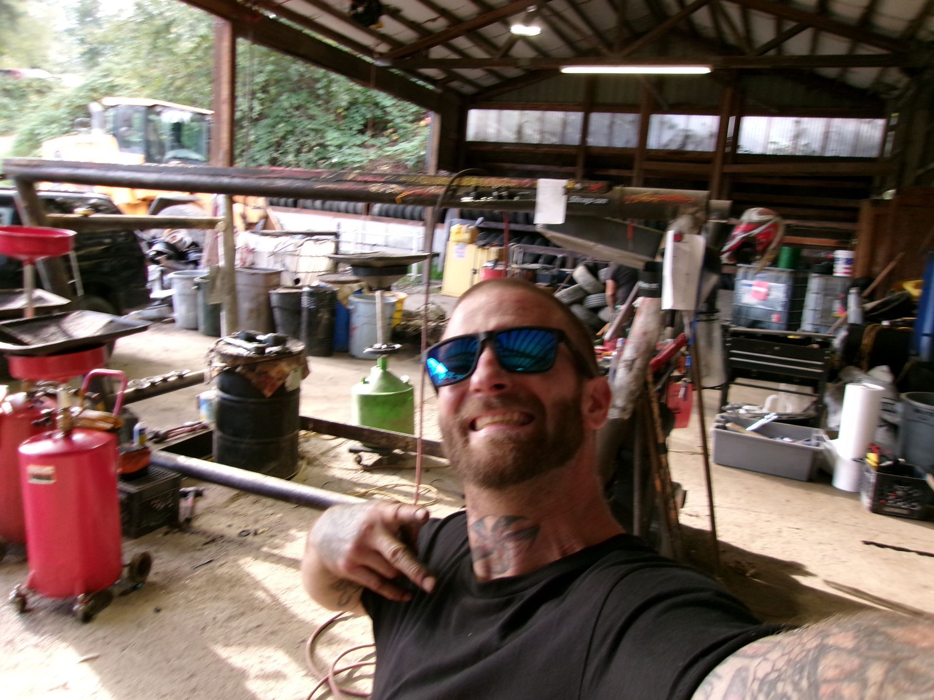 Man in sunglasses smiles at the camera in a cluttered workshop; a red air compressor is in view.