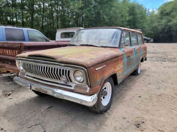 Rust-covered vintage Jeep Wagoneer in a salvage yard, with white wheels and blue paint showing through.