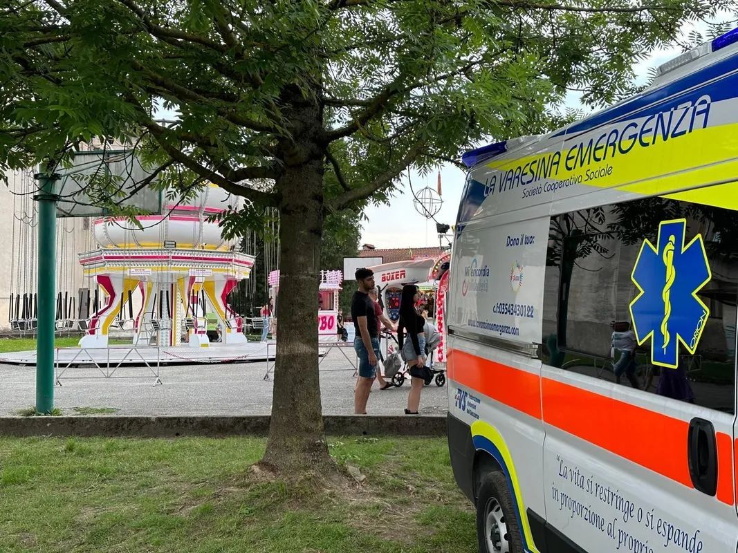Un'ambulanza è parcheggiata vicino a un albero, nei pressi di un padiglione colorato, e due persone camminano nelle vicinanze La Varesina Emergenza Varese