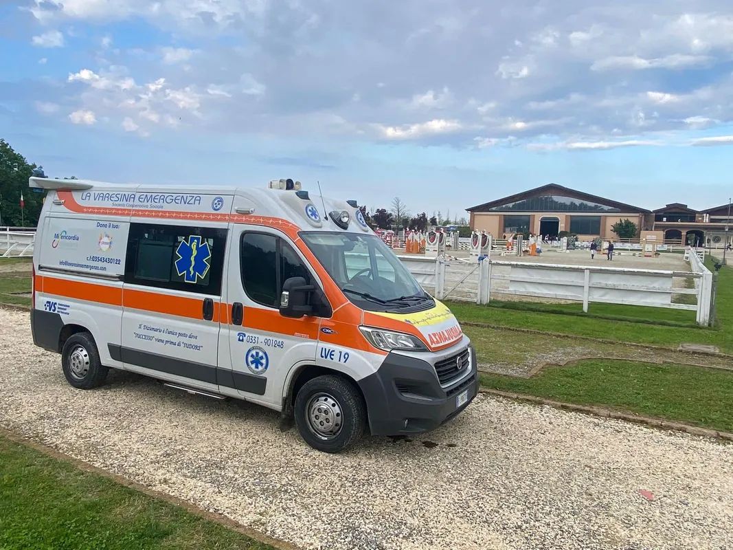 Ambulanza parcheggiata su ghiaia vicino a un campo recintato sotto un cielo nuvoloso La Varesina Emergenza Varese