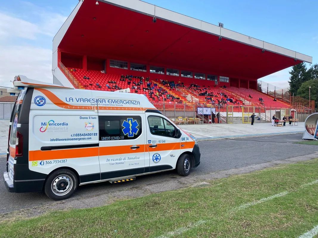 Un furgone ambulanza bianco è parcheggiato accanto a una tribuna rossa dello stadio, dove sono seduti gli spettatori La Varesina Emergenza Varese