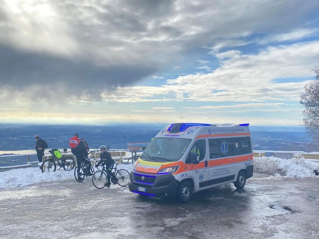 Ciclisti e un'ambulanza su una strada di montagna innevata sotto un cielo nuvoloso, con lo sguardo rivolto verso le colline in lontananza La Varesina Emergenza Varese