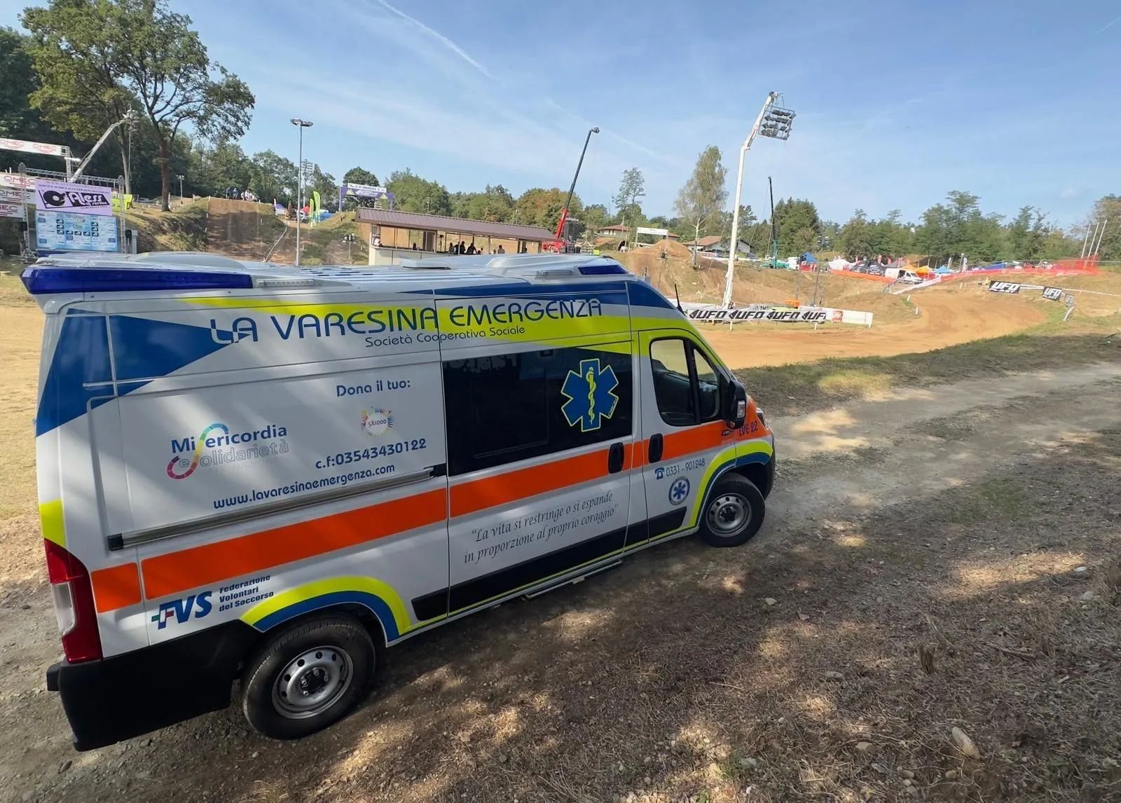 Furgone di emergenza bianco con strisce blu e arancioni parcheggiato su un campo sterrato sotto un cielo sereno.