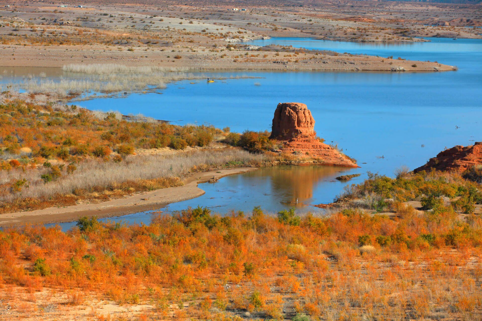 A lake with blue water, sandy shores, and a red rock formation, surrounded by orange and brown foliage under a clear sky.