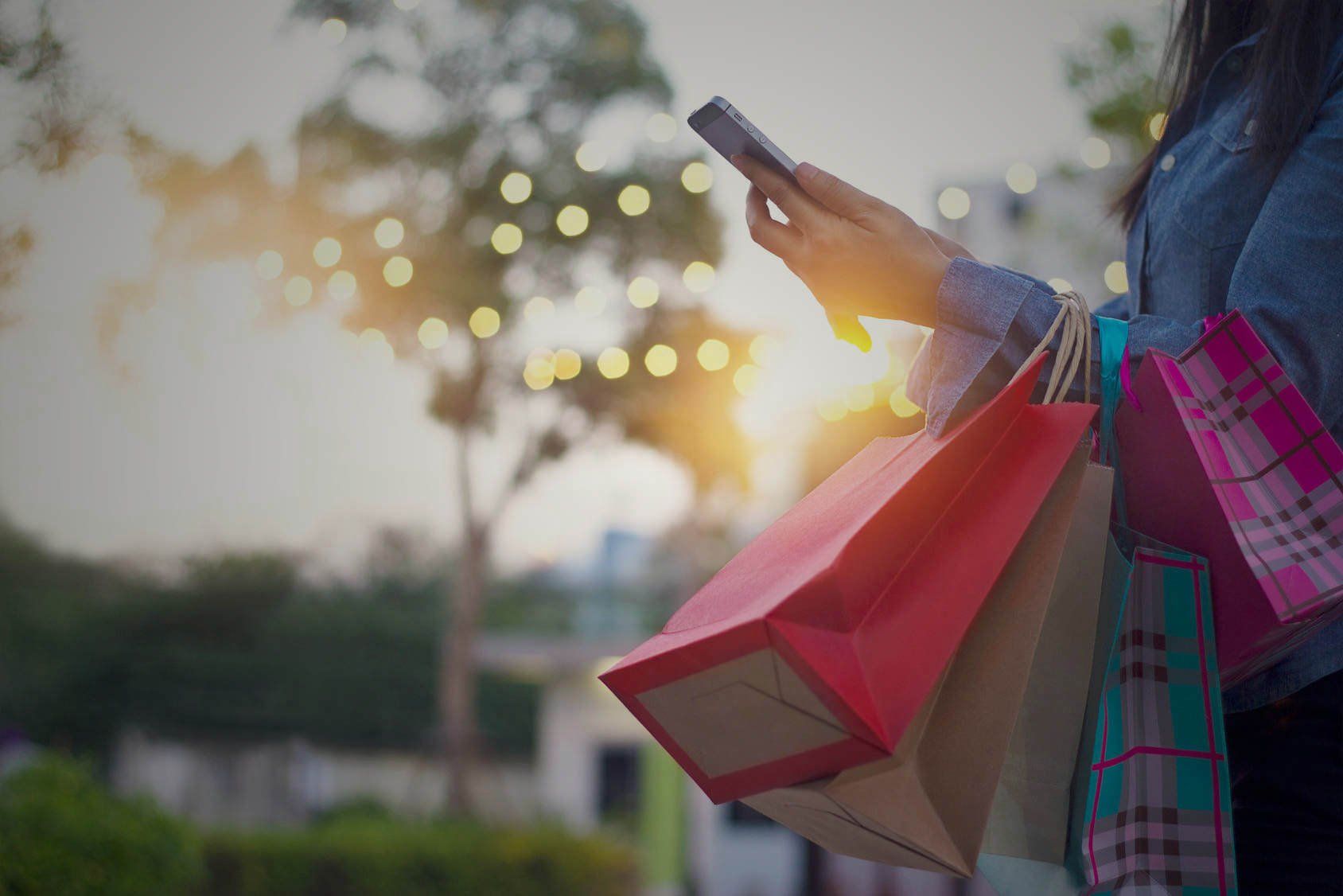 Woman holding shopping bags, using phone outdoors with warm lighting and trees in background.