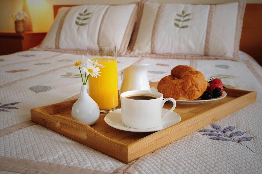 Breakfast tray on a bed with coffee, orange juice, croissant, and flowers.