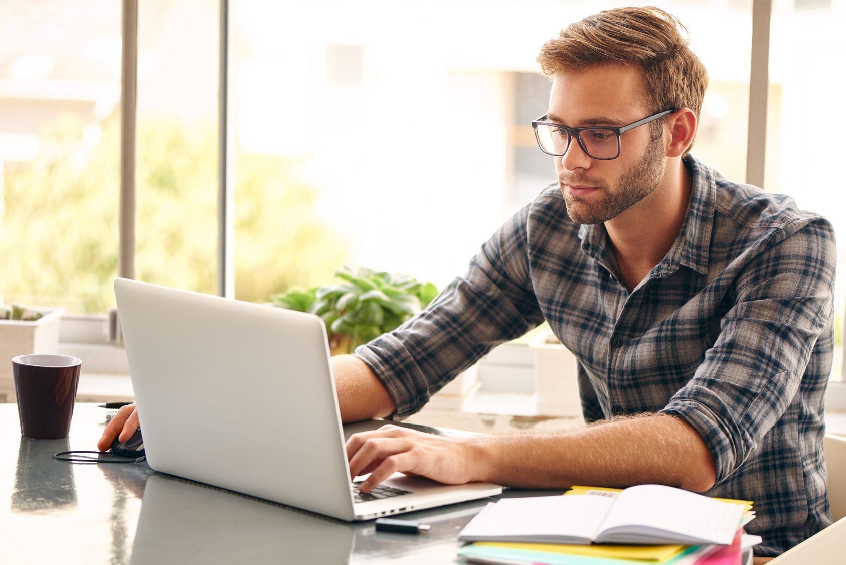 Man working on a laptop at a desk near a window, wearing glasses and a plaid shirt.