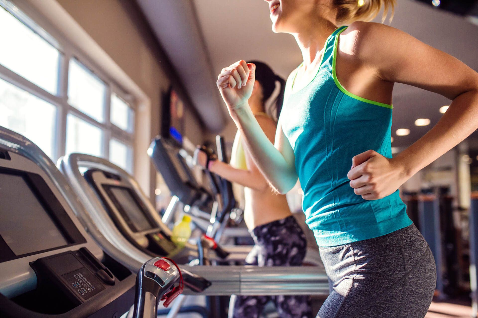 Two women running on treadmills in a gym near a window. One wears a teal tank top and gray leggings.