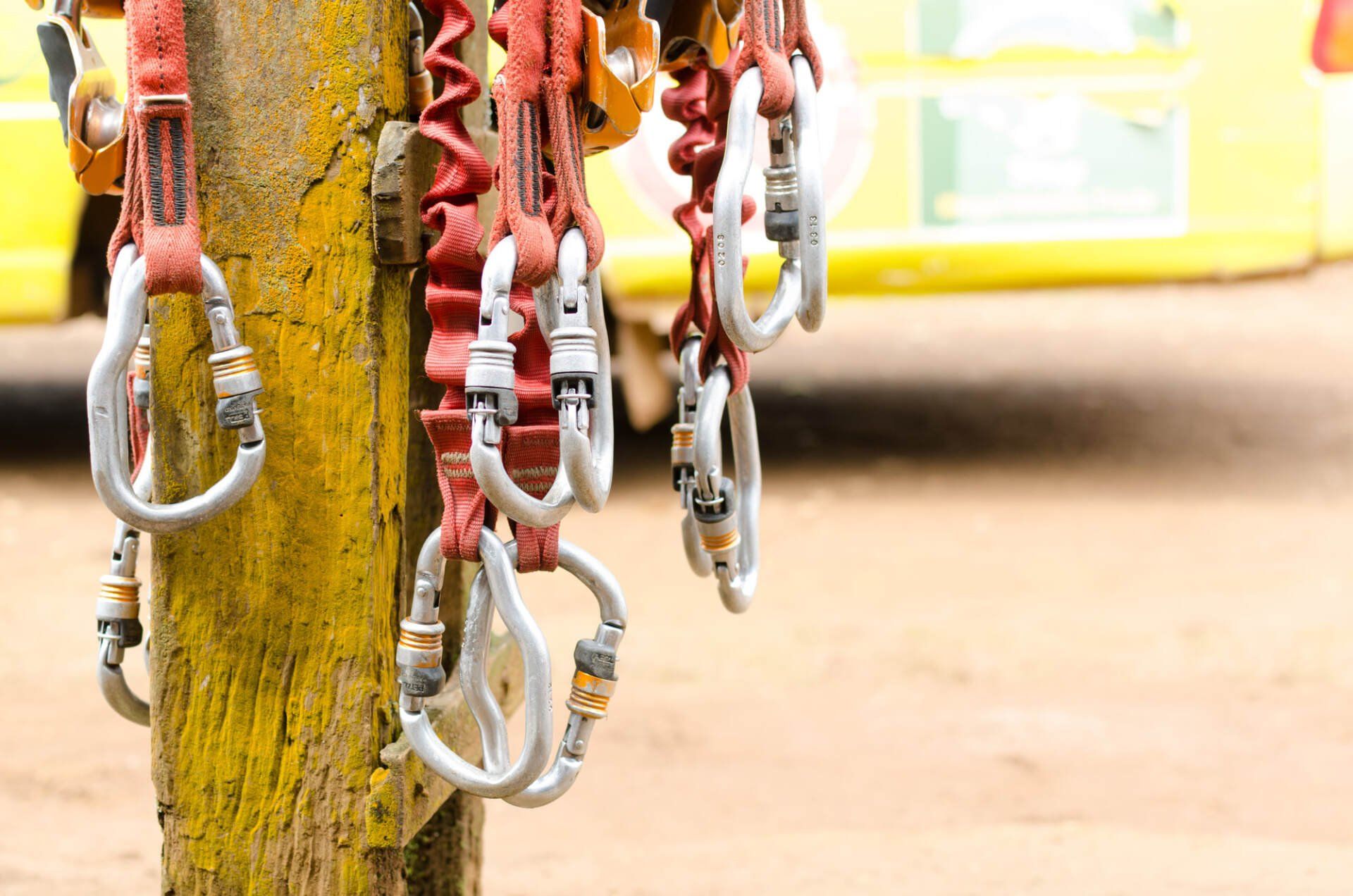 Climbing harnesses with carabiners hanging on a yellow post, yellow vehicle in background.